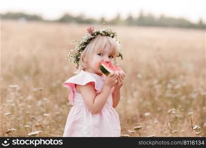 picnic with family. cute little girl eating big piece of watermelon on the grass in summertime in the park. Adorable child wearing in flowers wreath on head and pink dress. picnic with family. cute little girl eating big piece of watermelon on the grass in summertime in the park. Adorable child wearing in flowers wreath on head and pink dress.