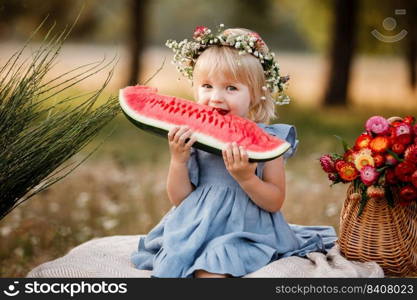 picnic with family. cute little girl eating big piece of watermelon on straw stack in summertime in the park. Adorable child wearing in flowers wreath on head and blue dress. picnic with family. cute little girl eating big piece of watermelon on straw stack in summertime in the park. Adorable child wearing in flowers wreath on head and blue dress.