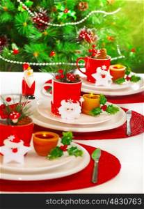 Picture of festive table setting, red cup for tea full of different Christmastime decoration, small Santa Clause decor with yellow candle on white white plate on Christmas tree background