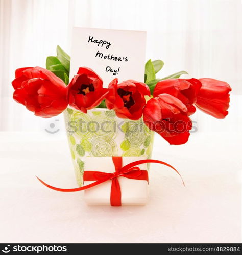 Picture of fresh red tulip flowers in beautiful vase with paper greeting postcard on the table, little white gift box with ribbon, home interior, happy mothers day, festive still life, love concept