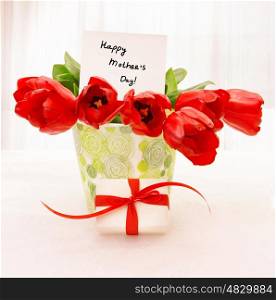 Picture of fresh red tulip flowers in beautiful vase with paper greeting postcard on the table, little white gift box with ribbon, home interior, happy mothers day, festive still life, love concept