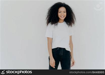 Picture of good looking woman with gentle smile, stands satisfied, has sincere carefree expression, relaxed casual mood, dressed in casual tshirt and black jeans, isolated over white background