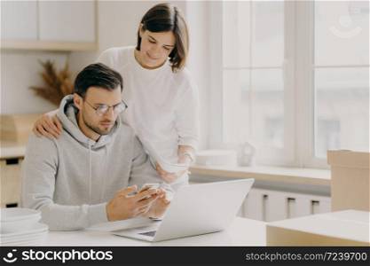 Picture of husband and wife study notification from bank, holds mobile phone and papers, work on laptop computer, pose in kitchen during relocation day, dressed in casual wear, save family money