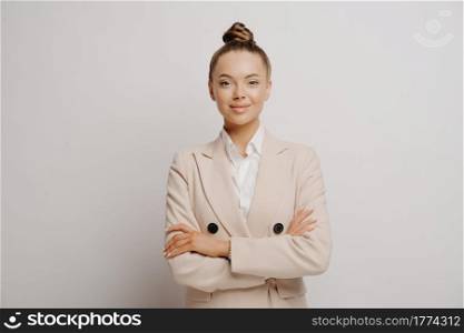 Picture of young beautiful woman manager in beige suit and white shirt with crossed arms, slightly smiling and looking straight forward while being succesfull and confident against to light wall. Succsesfull woman manager in beige suit with crossed arms