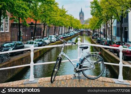 Picturesque Holland cityscape view - bicycle on bridge and canal with cars parked along and Oude Kerk old church in street of Delft, Netherlands. Bicycle on bridge and canal with cars parked along and Oude Kerk old church in Delft street