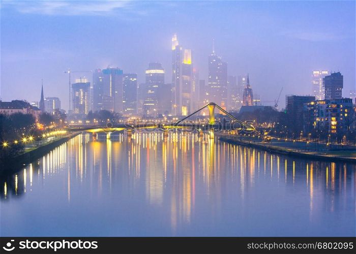 Picturesque view of foggy Frankfurt am Main skyline during evening blue hour with mirror reflections in the river, Germany