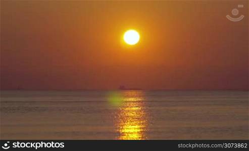 Picturesque view of sundown over the sea with cargo ship and sailboat in the distance, golden sun reflecting in still water