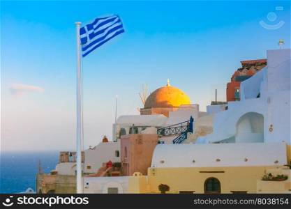 Picturesque view of white houses, church and Greek flag in the morning, Oia or Ia, island Santorini, Greece