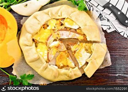 Pie of pumpkin, pear, soft cheese and walnuts on parchment, napkin and knife, parsley on a wooden board background from above