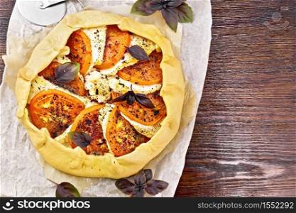 Pie with tomatoes, curd cheese and purple basil on parchment on a wooden board background from above