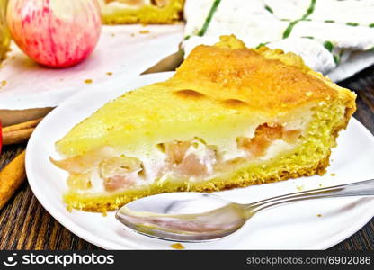 Piece of apple pie with sour cream sauce in a white plate, cinnamon, fruit and napkin on a background of a dark wooden board