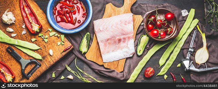 Piece of fish fillets on rustic cutting board with seasonal vegetables cooking ingredients and tomatoes sauce, dark styled background, top view, banner