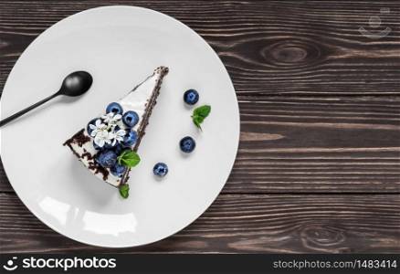 Piece of homemade bird cherry cake with sour cream, decorated with blueberries and mint leaves on a white plate. Siberian traditional cake, top view with copy space, dark wooden background. Piece of homemade bird cherry cake with sour cream, decorated with blueberries and mint leaves on a white plate. dark wooden background