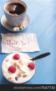 Piece of paper with thank you text and a smiley face, a healthy breakfast with yogurt and fruits, a cup of arabic coffee, on a blue background in the morning light.