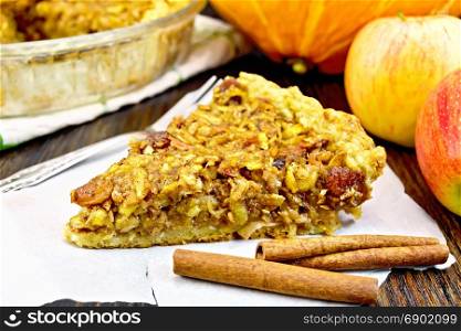Piece of pie with apples, pumpkin, raisins and nuts on parchment, cinnamon on the background of a wooden board
