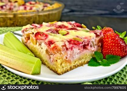 Piece of pie with strawberries, rhubarb and cream sauce, fork, strawberries, rhubarb stalks, mint in white plate on a green towel on a dark wooden board