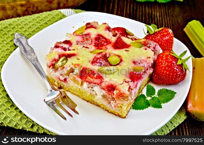 Piece of pie with strawberries, rhubarb and cream sauce, fork, strawberry, mint in white plate on a green towel, rhubarb stalks on a wooden boards background