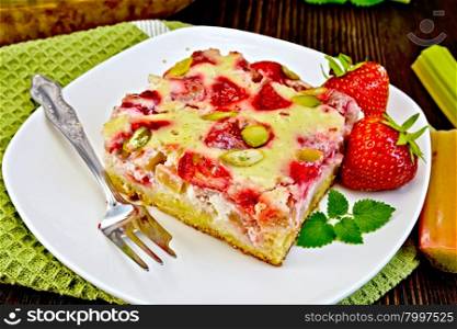 Piece of pie with strawberries, rhubarb and cream sauce, fork, strawberry, mint in white plate on a green towel, rhubarb stalks on a wooden boards background