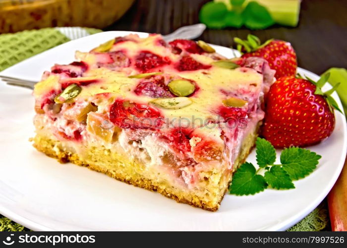 Piece of pie with strawberries, rhubarb and cream sauce, fork, strawberry, mint in white plate on a towel, rhubarb stalks on a wooden boards background