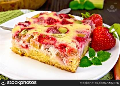 Piece of pie with strawberries, rhubarb and cream sauce, fork, strawberry, mint in white plate on a towel, rhubarb stalks on a wooden boards background