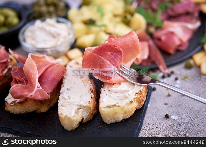 piece of prosciutto ham on a fork with traditional antipasto meat plate on background.. piece of prosciutto ham on a fork with traditional antipasto meat plate on background