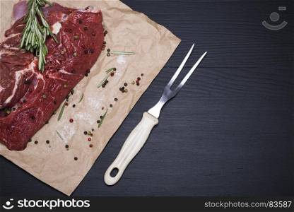 piece of raw beef on brown sheet of paper and fork, black wooden background, empty space on the right