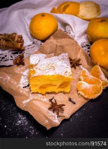 piece of tangerine cake on brown kraft paper and fresh tangerines, black wooden table, top view