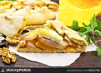 Piece pie of pumpkin, pear, soft cheese and walnuts on parchment, parsley on a dark wooden board