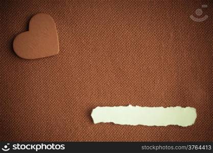 Piece scrap of white paper blank copy space and heart symbol of love on brown fabric textile material background. Valentines day. Sepia tone.
