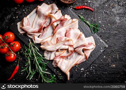Pieces of bacon on a stone board with tomatoes and rosemary. On a black background. High quality photo. Pieces of bacon on a stone board with tomatoes and rosemary.