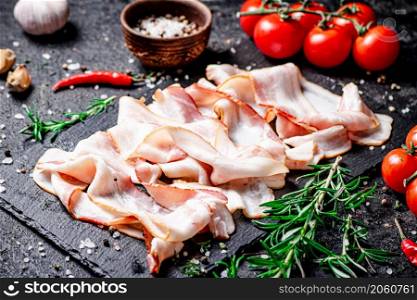 Pieces of bacon on a stone board with tomatoes and rosemary. On a black background. High quality photo. Pieces of bacon on a stone board with tomatoes and rosemary.