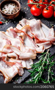 Pieces of bacon on a stone board with tomatoes and rosemary. On a black background. High quality photo. Pieces of bacon on a stone board with tomatoes and rosemary.