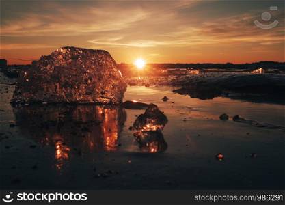 Pieces of beautiful transparent ice in pink and amber colors as a jewels shining on a frozen seashore in the sunset