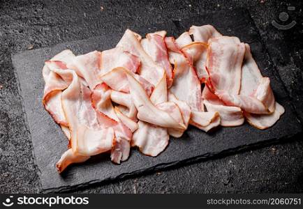 Pieces of fresh bacon on a stone board. On a black background. High quality photo. Pieces of fresh bacon on a stone board.