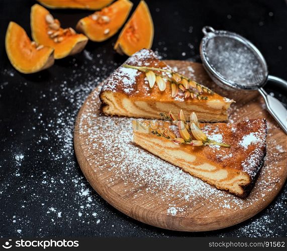 pieces of pumpkin pie on a round wooden board are sprinkled with sugar powder, a black wooden background with pieces of fresh pumpkin