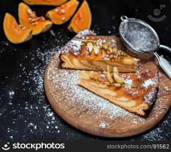 pieces of pumpkin pie on a round wooden board are sprinkled with sugar powder, a black wooden background with pieces of fresh pumpkin