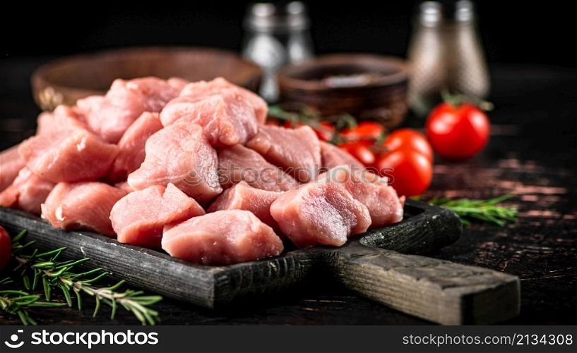 Pieces of raw pork on a cutting board with spices, tomatoes and rosemary. On a rustic dark background. High quality photo. Pieces of raw pork on a cutting board with spices, tomatoes and rosemary.