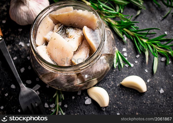 Pieces of salted herring in a glass jar with rosemary and garlic. On a black background. High quality photo. Pieces of salted herring in a glass jar with rosemary and garlic.