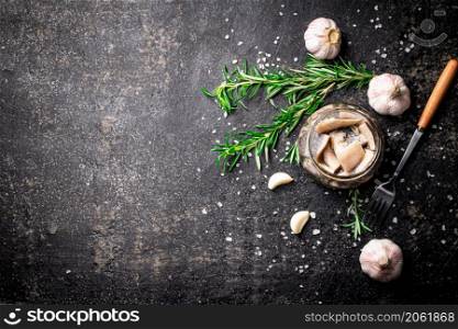 Pieces of salted herring in a glass jar with rosemary and garlic. On a black background. High quality photo. Pieces of salted herring in a glass jar with rosemary and garlic.