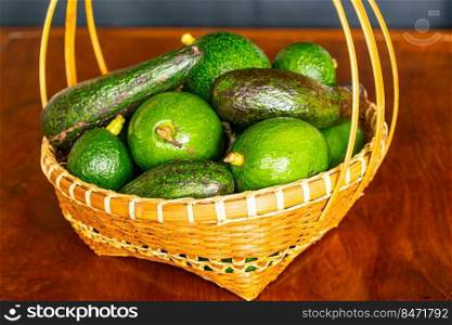 Pile of different varieties of fresh green avocado in wicker bamboo basket on wooden table.