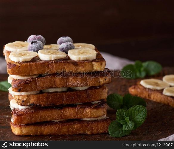 pile of square fried bread slices with chocolate and banana slices on a brown wooden board, French toast for breakfast
