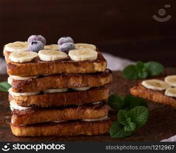 pile of square fried bread slices with chocolate and banana slices on a brown wooden board, French toast for breakfast
