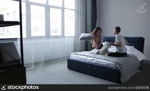 Pillow fight. Beautiful young couple fighting with pillows in bed and smiling. Excited joyful couple in love spending great time together having fun in bedroom in the morning. Slow motion. Steadicam stabilized shot.
