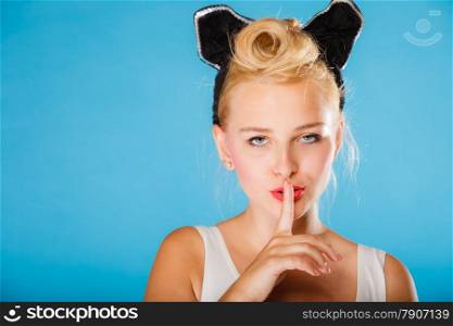 Pin up and retro style. Young smiling woman with black ears on head and finger silence sign symbol on blue background. Studio shot.