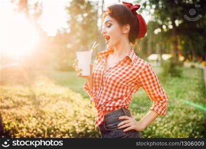 Pin up girl holds cardboard cup with a straw, city park on background. Vintage american fashion. Attractive woman in pinup style