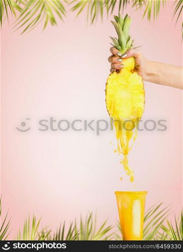 Pineapple juice concept. Female hand holding half of pineapple and presses juice into glass at pink background with tropical palm leaves. Summer beverages splash