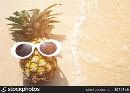 Pineapple with sunglasses sunbathe sunshine on tropical beach with a wave&rsquo;s edge foaming gently beside them. Fashion Hipster summer Minimal style. vacation travel holiday on ocean beach concept.