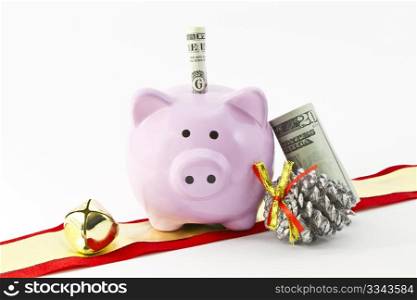 Pink bank shaped like a pig is placed with 2 dollar bill at top and 20 dollar bill leaning on it next to silver pine cone, red and gold Christmas ribbon, and gold Christmas bells; copy space; nobody;