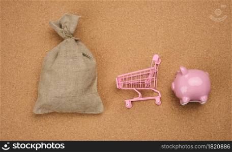 pink ceramic piggy bank, miniature shopping cart and full canvas bag on brown background, top view.