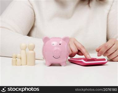 pink ceramic piggy bank, wooden family figurines. Woman sitting at table and counting on calculator, budget planning concept